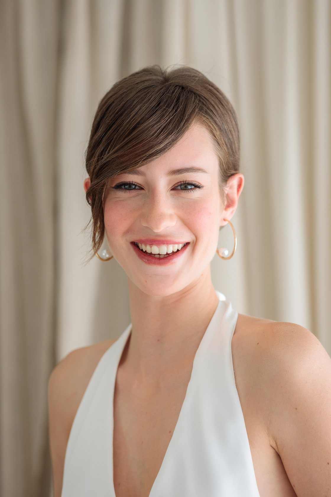Finished look: smiling model with hair swept up and side bangs, white halter dress, gold hoop earrings