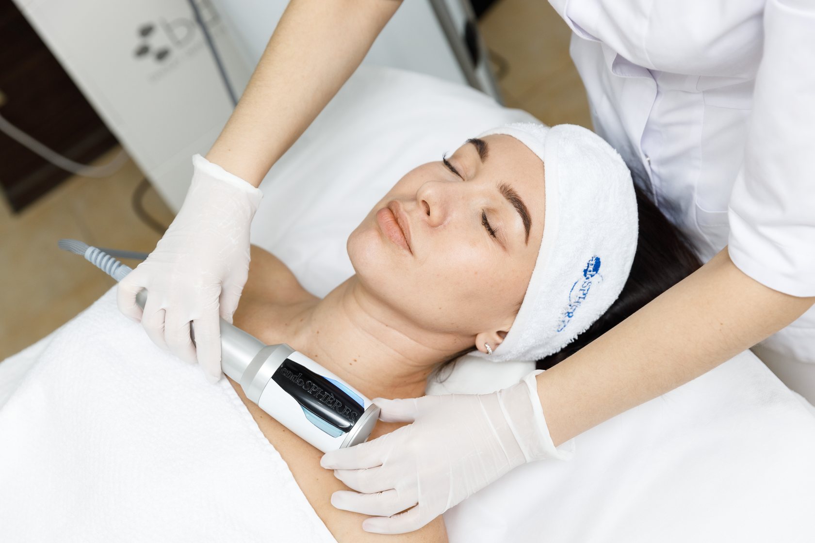 Client with eyes closed receiving a device treatment on her neck, practitioner in white gloves